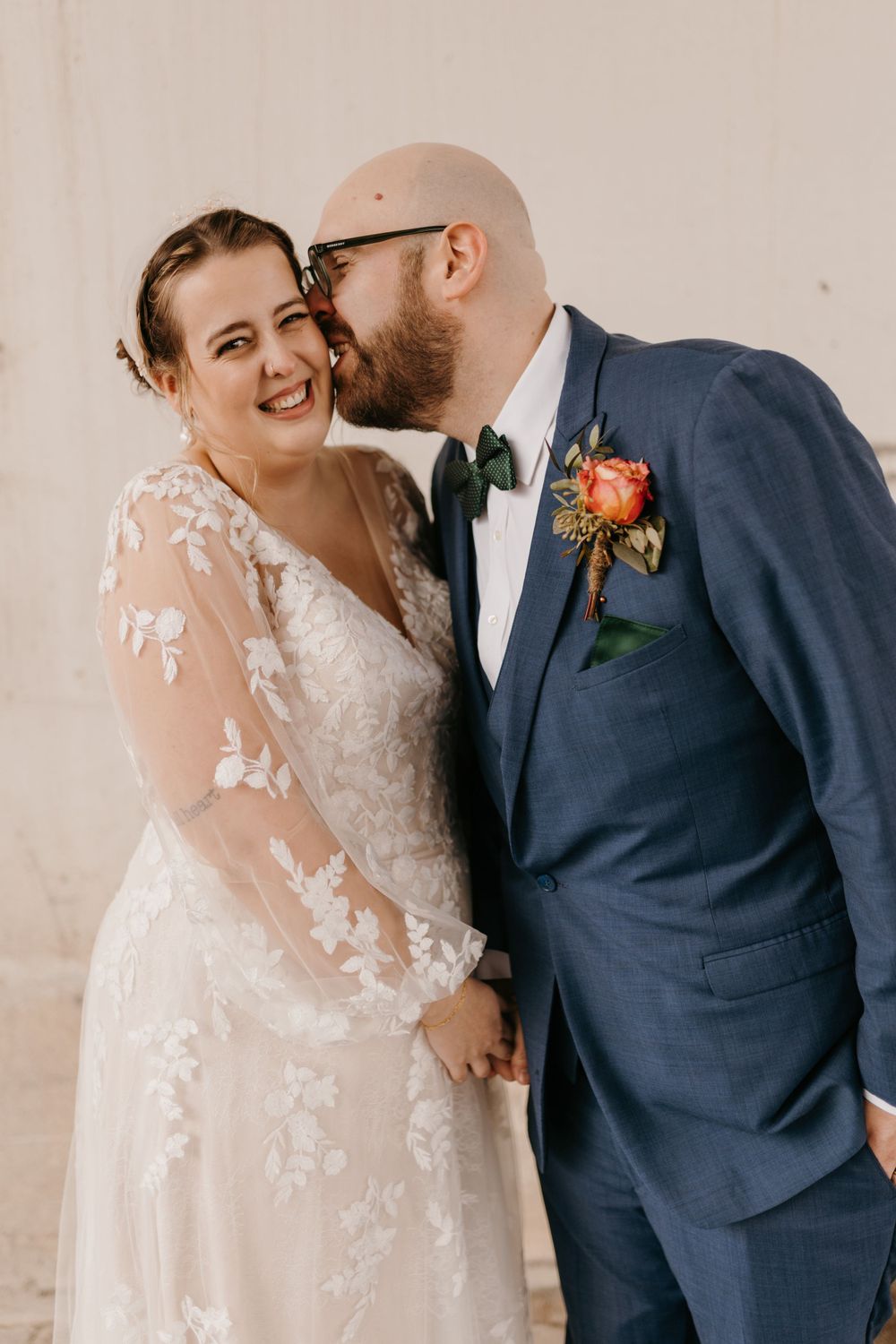 Groom in navy suit kissing bride on cheek as she smiles in lace wedding gown.