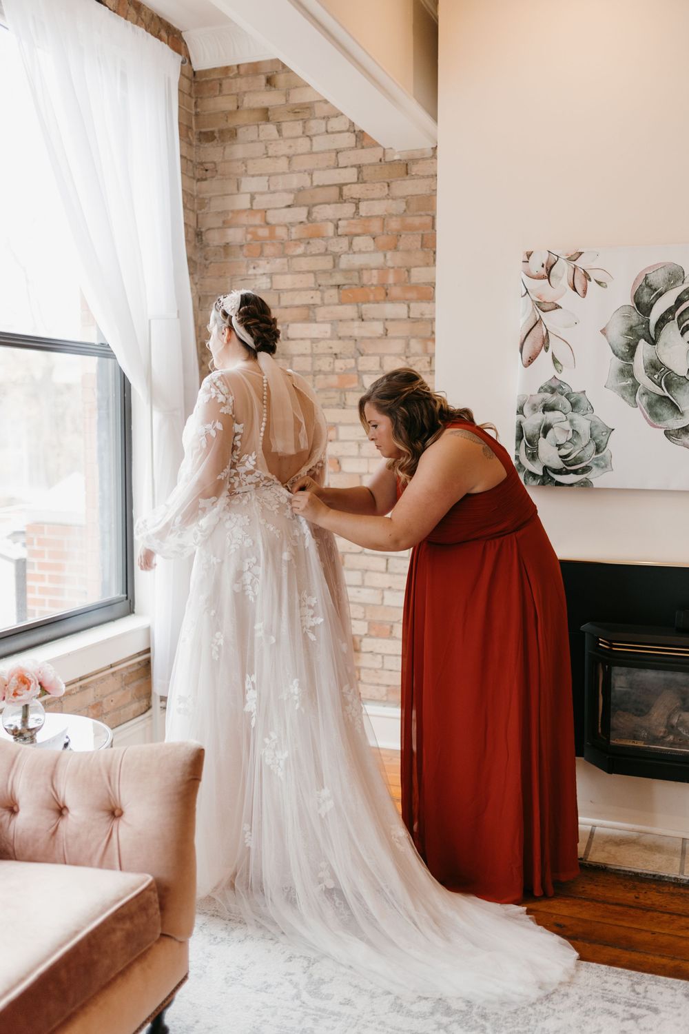 Bridesmaid in burgundy dress fastening back of bride's lace wedding gown by window.