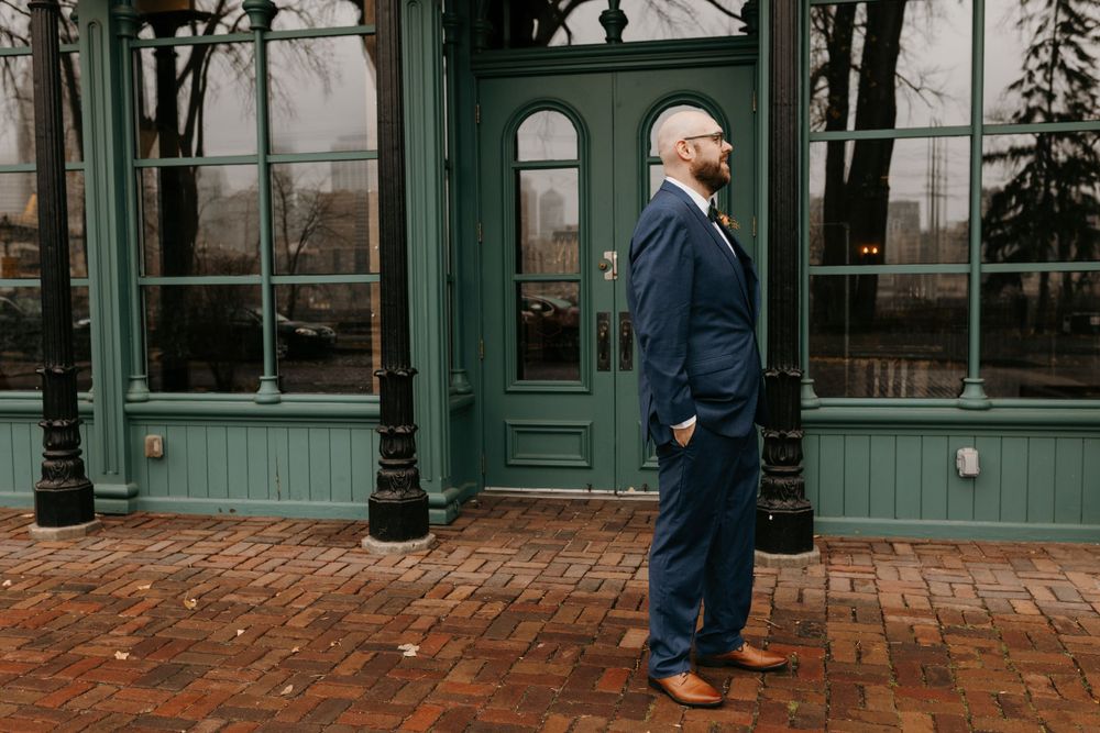 Groom in navy blue suit standing proudly before green vintage building on brick pathway.