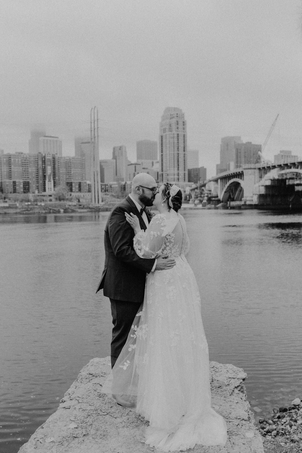 Black and white photo of newlyweds sharing a kiss by the river with city skyline behind.