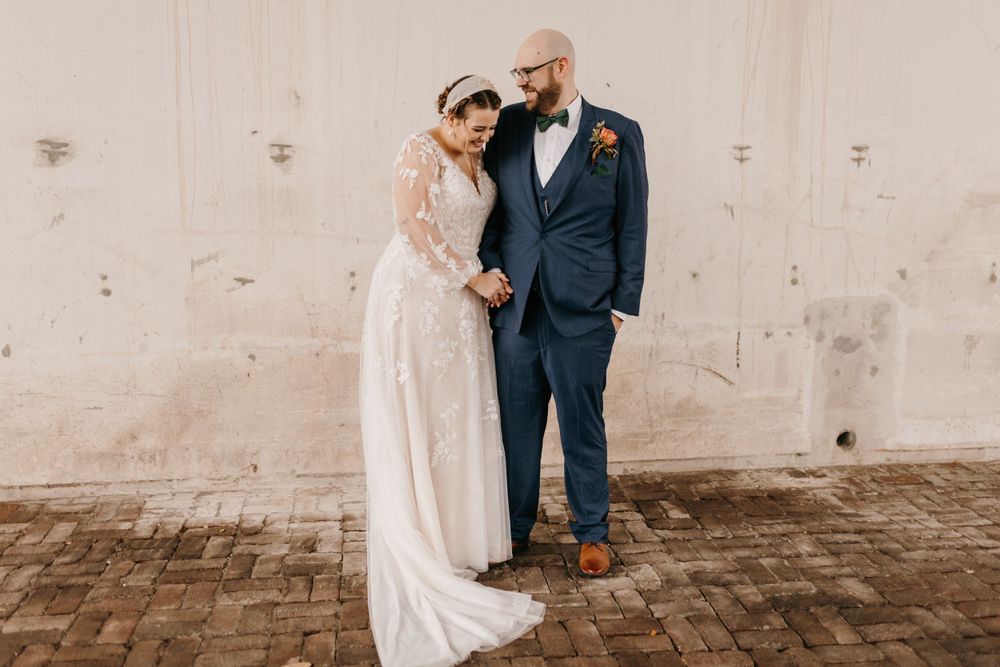 Bride leaning into groom while standing together against weathered white wall on their wedding day.