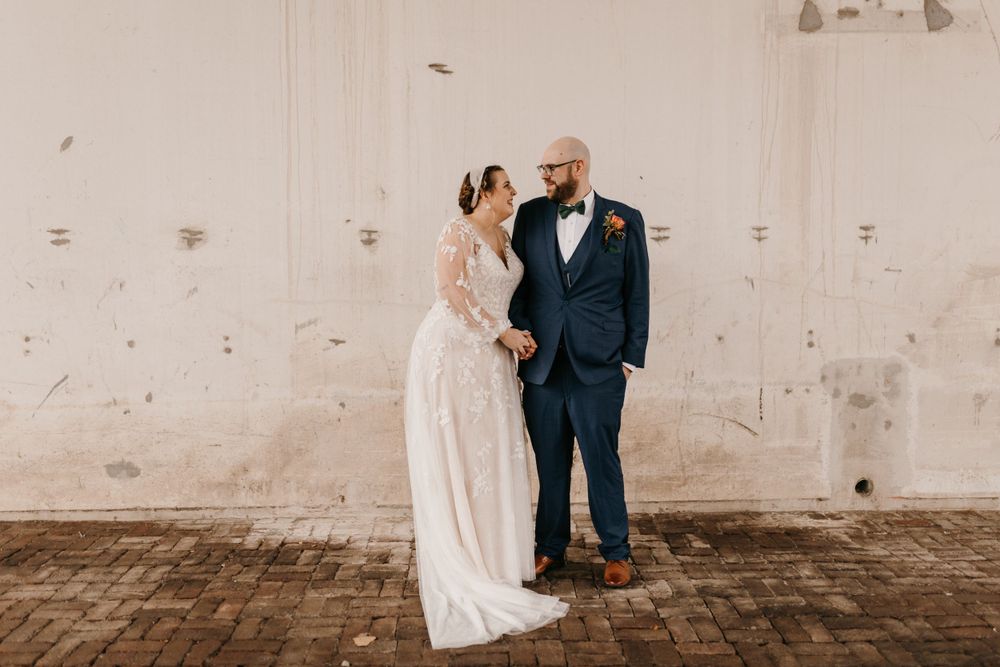 Newlyweds smiling and holding hands in front of rustic white textured wall on their wedding day.