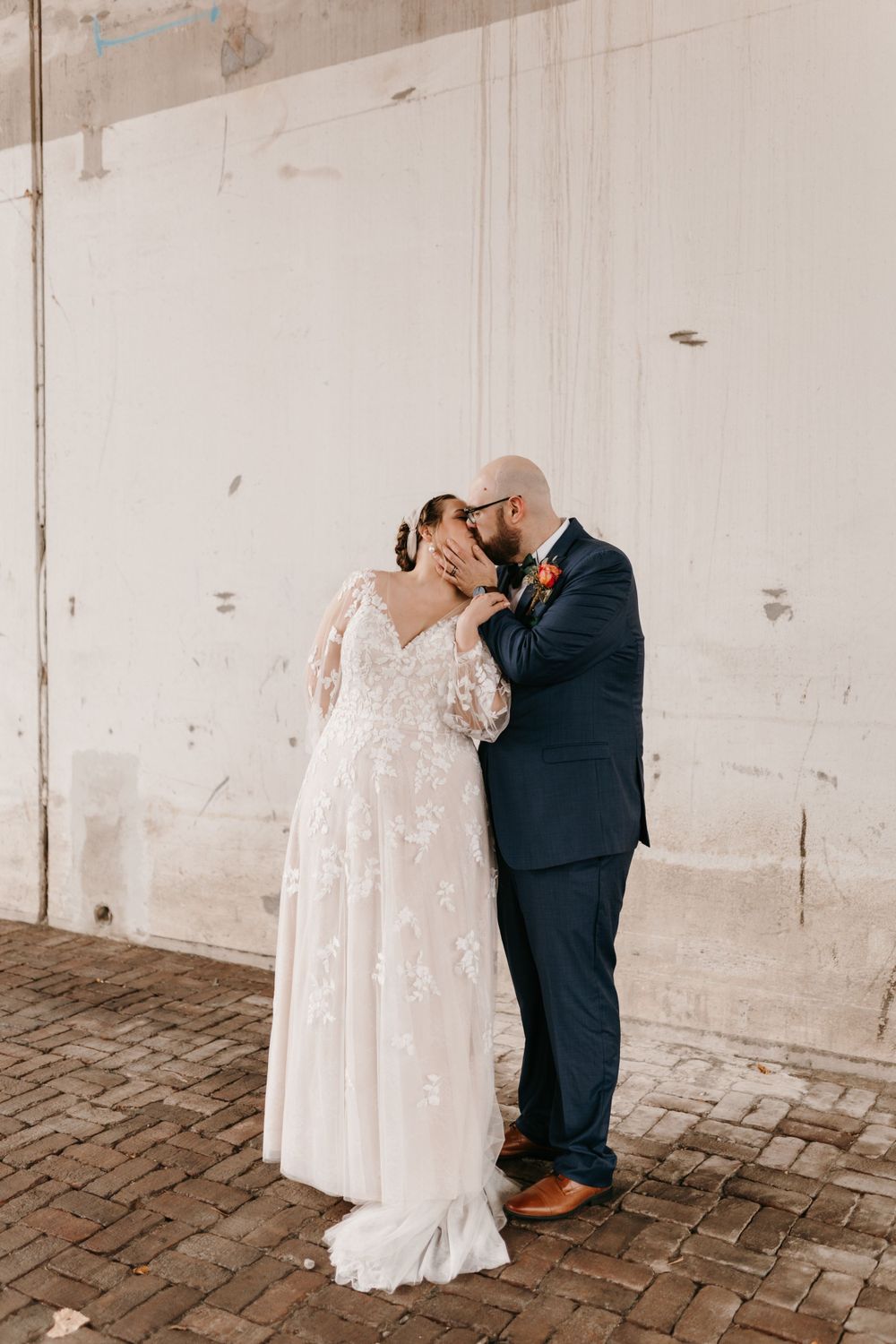 Bride and groom sharing a romantic kiss against white wall on brick path.