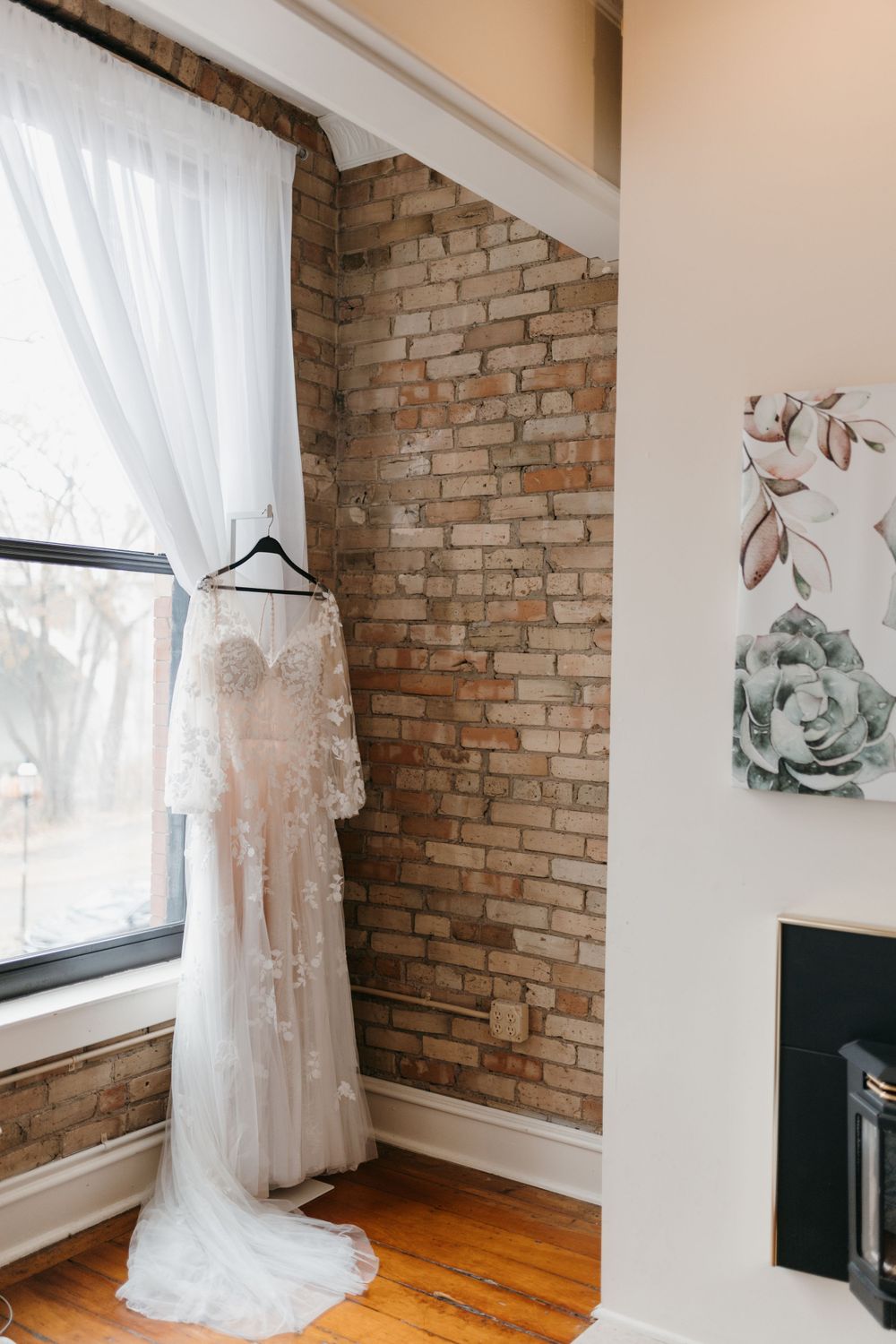 Lace bridal gown hanging on a black hanger against a rustic exposed brick wall.
