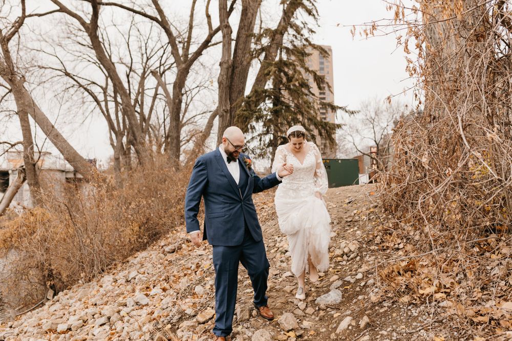 Groom helping bride walk up a rocky hillside surrounded by bare autumn trees.