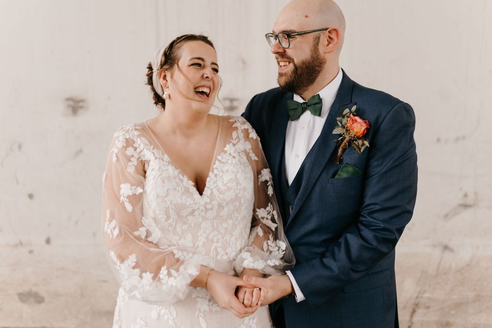 Bride laughing joyfully while holding hands with groom in navy suit outdoors.