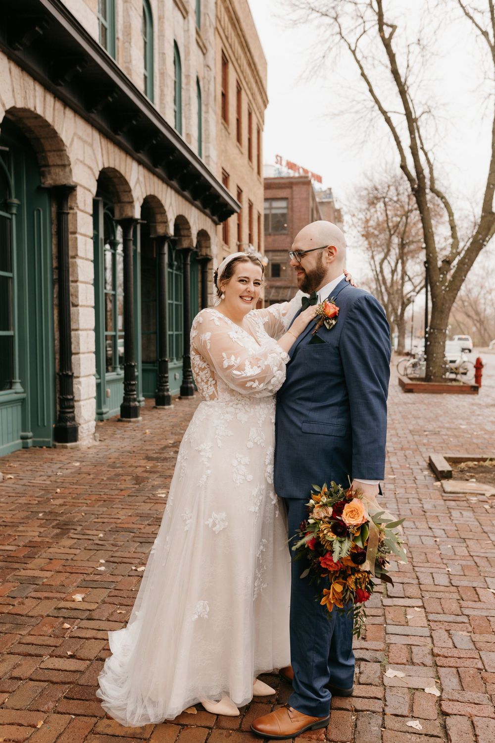 Bride in lace gown adjusting groom's boutonniere while smiling on brick sidewalk in historic district.