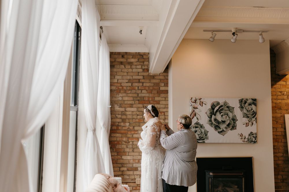 Bridesmaid helping bride with veil near sheer curtains and exposed brick wall interior.