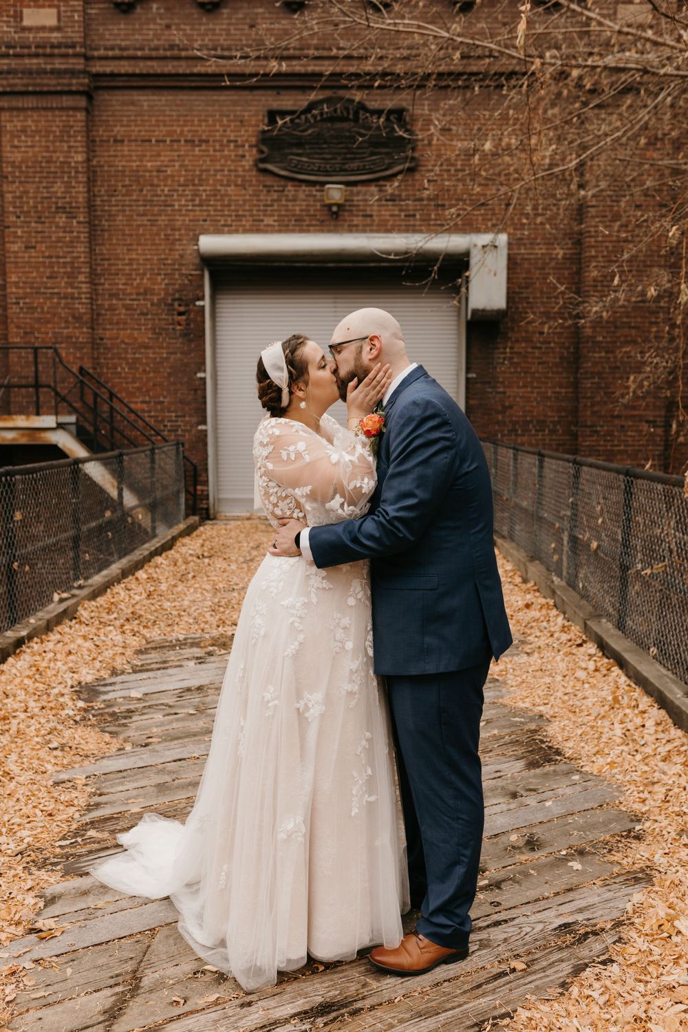 Bride and groom share a romantic kiss on a leaf-covered walkway outside brick venue.