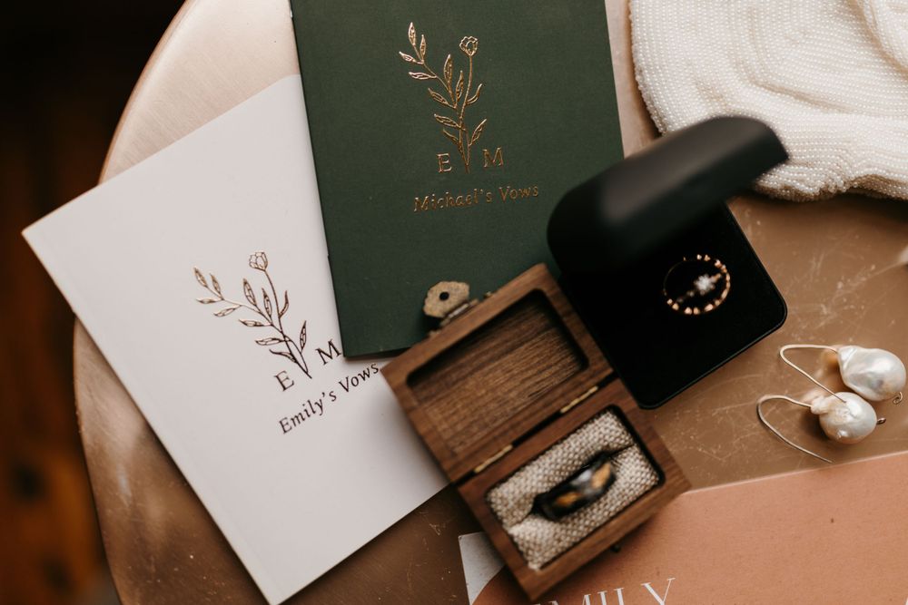 Elegant wedding flat lay featuring vow books, ring boxes, floral earrings, and invitation suite.