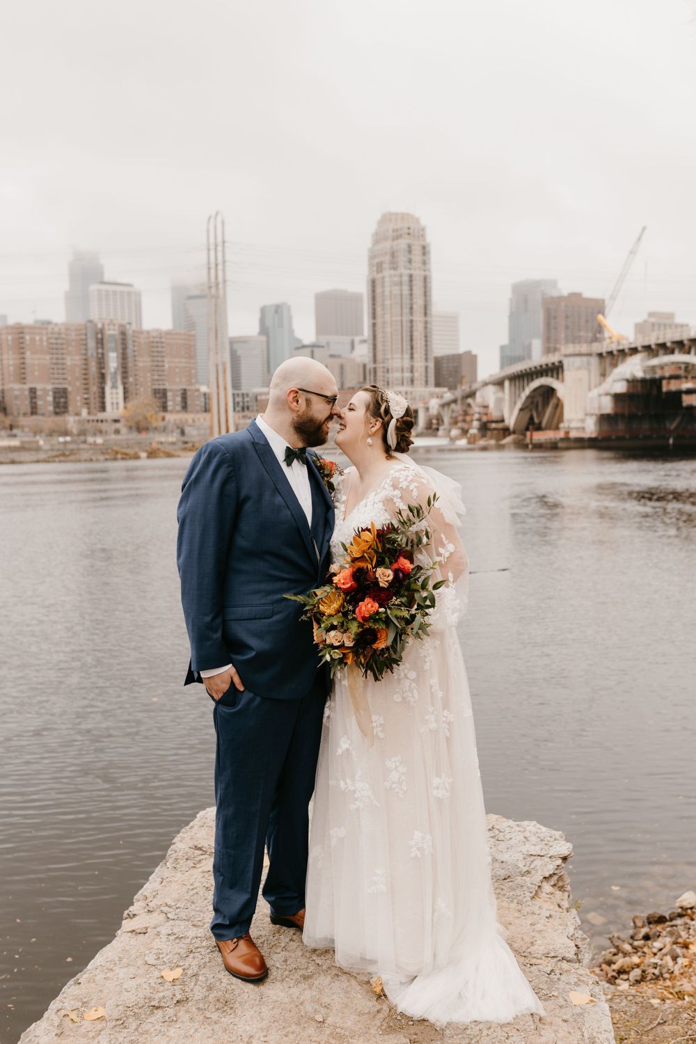 Bride and groom nearly kissing on riverbank rocks with colorful bouquet and city skyline.