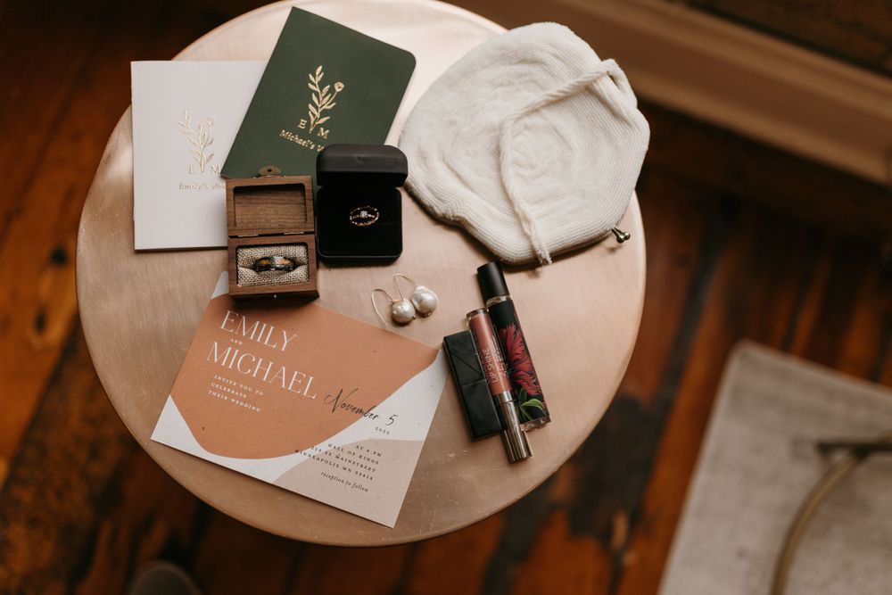 Flat lay of wedding details including invitation, rings, earrings, and vow books on a round table.
