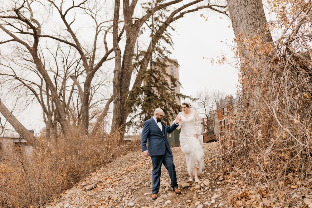 Bride and groom walking hand in hand through a leaf-covered autumn landscape with bare trees.