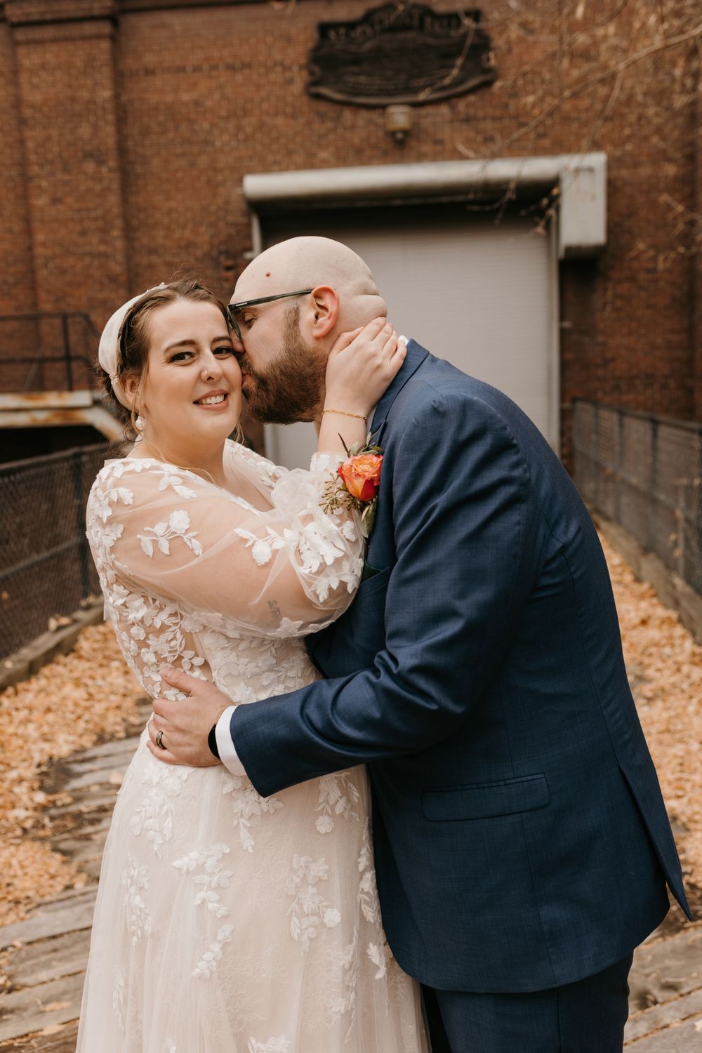 Groom kisses bride on the cheek as she smiles at camera in front of brick building.