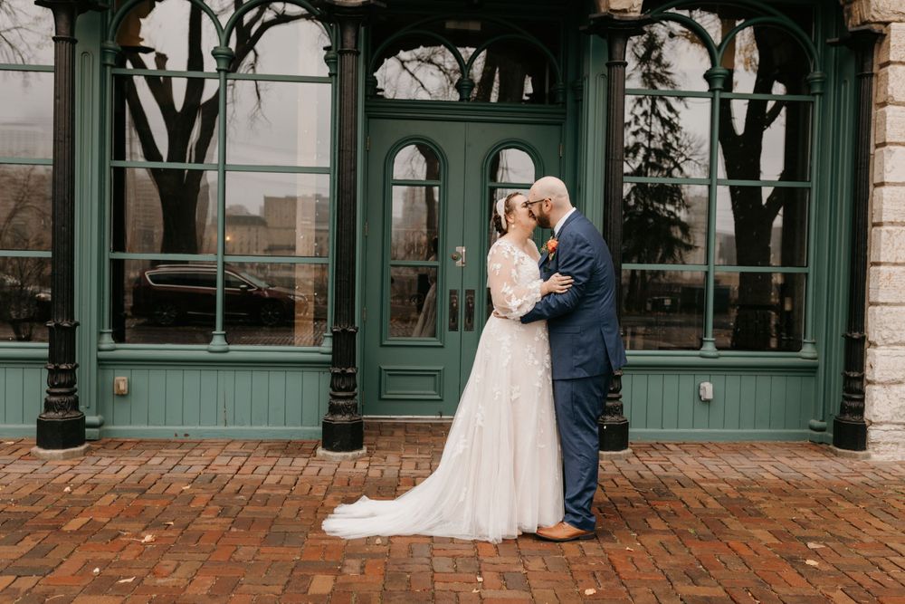 Newlyweds sharing romantic kiss outside elegant green vintage building on brick pathway.