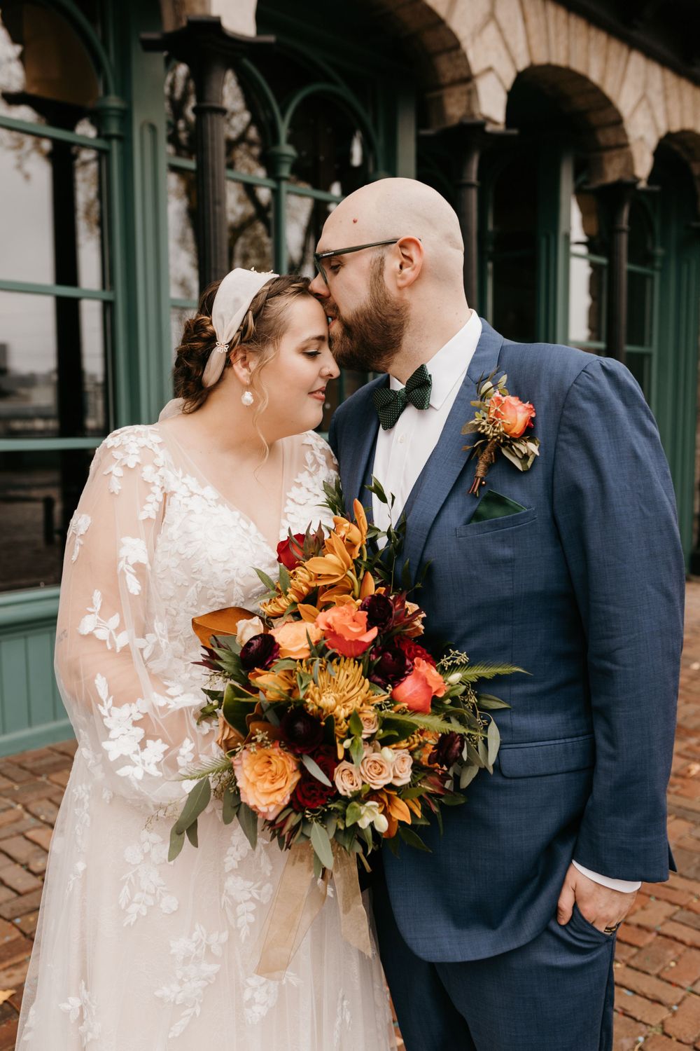 Groom kissing bride on forehead as she holds stunning autumn wedding bouquet outdoors.