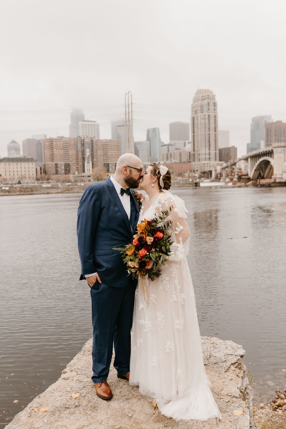 Bride and groom kissing on rocky riverbank holding vibrant fall bouquet with city skyline.