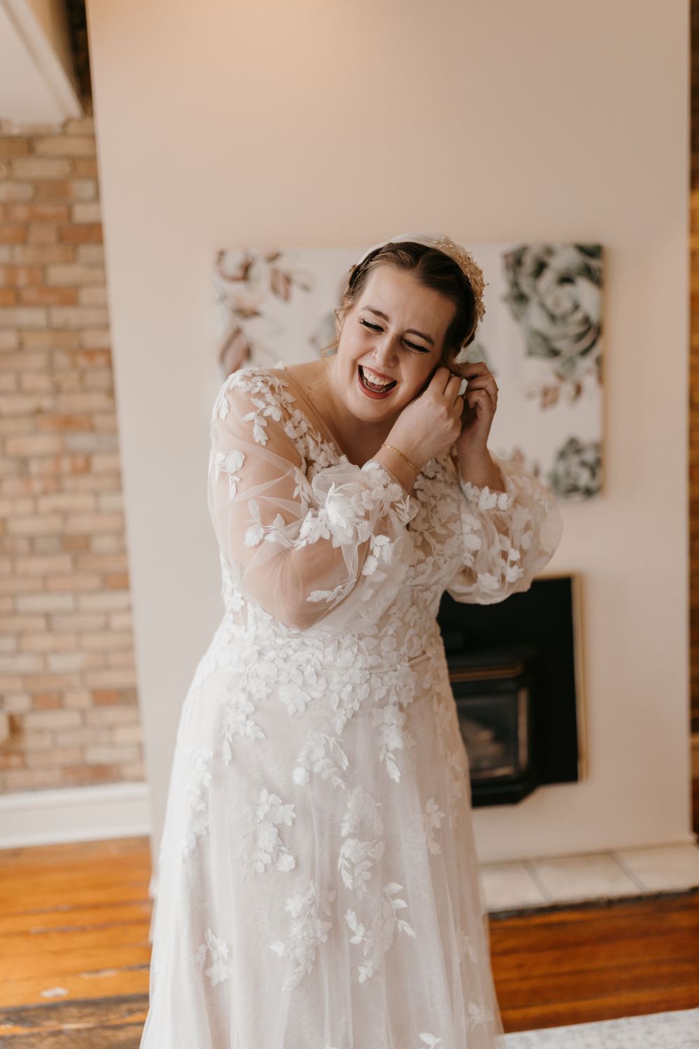 Laughing bride in floral lace wedding dress adjusting earring near exposed brick fireplace.