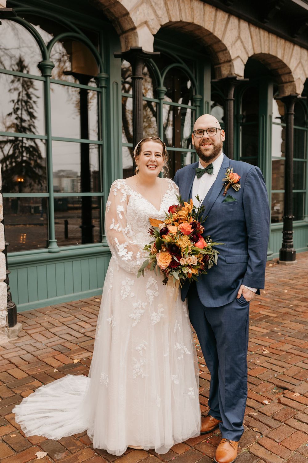 Smiling bride and groom posing with vibrant autumn bouquet outside green historic venue.