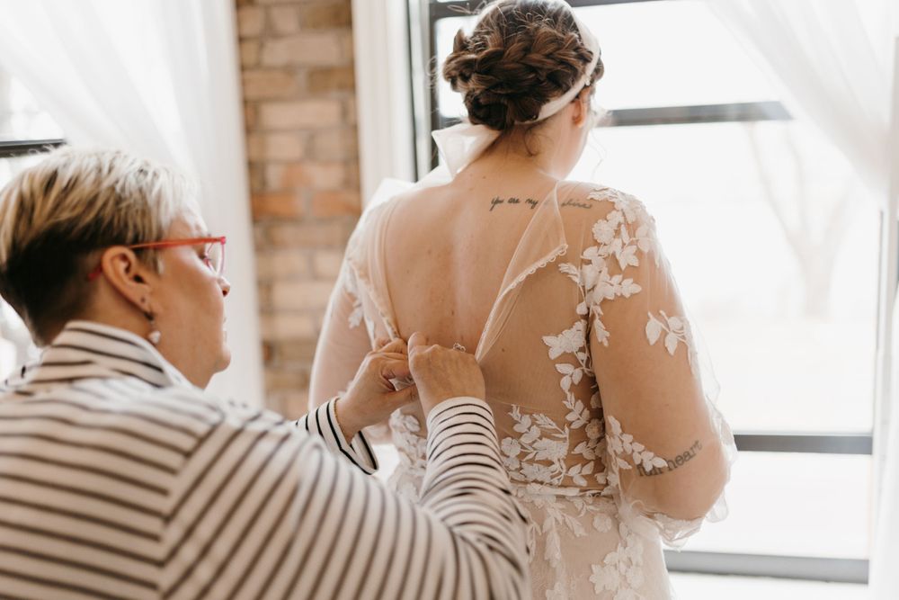 Woman in striped shirt carefully buttoning the back of a floral lace bridal gown.