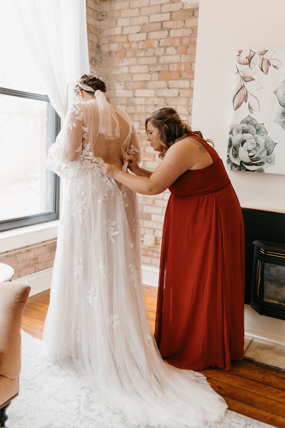 Bridesmaid in red gown buttoning bride's floral lace wedding dress near brick wall.