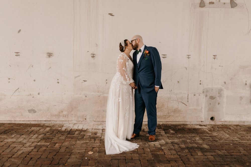 Bride and groom sharing a tender kiss against a weathered white wall on brick pavement.