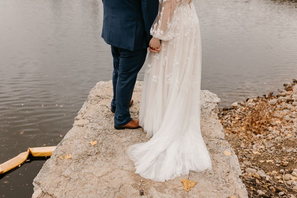 Close-up of bride in lace gown and groom in navy suit standing on rocky riverbank.