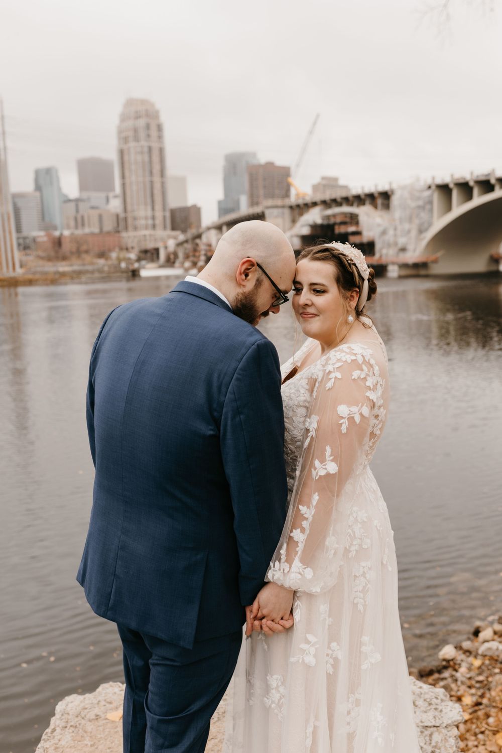 Wedding couple holds hands tenderly by Mississippi River with city skyline visible behind them.