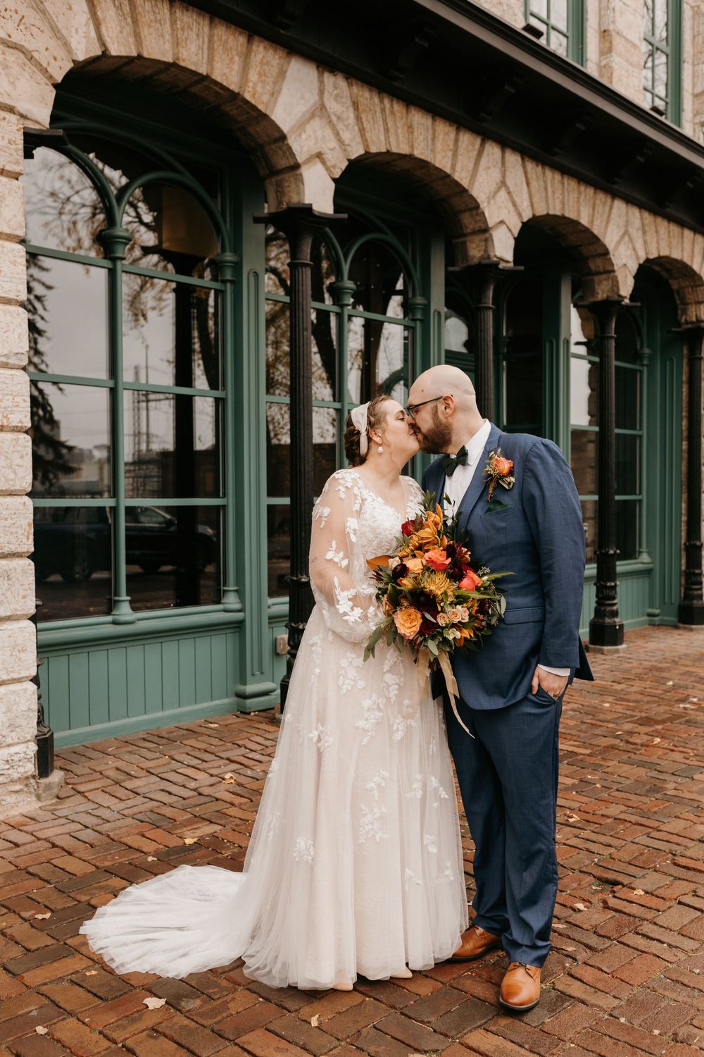 Bride and groom sharing romantic kiss with colorful fall bouquet outside green building.