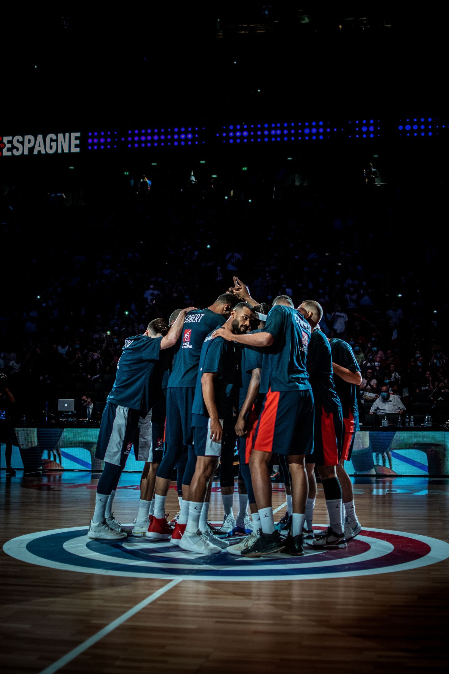 Basketball team huddles together on court during game with blue lighting and Espagne sign visible.
