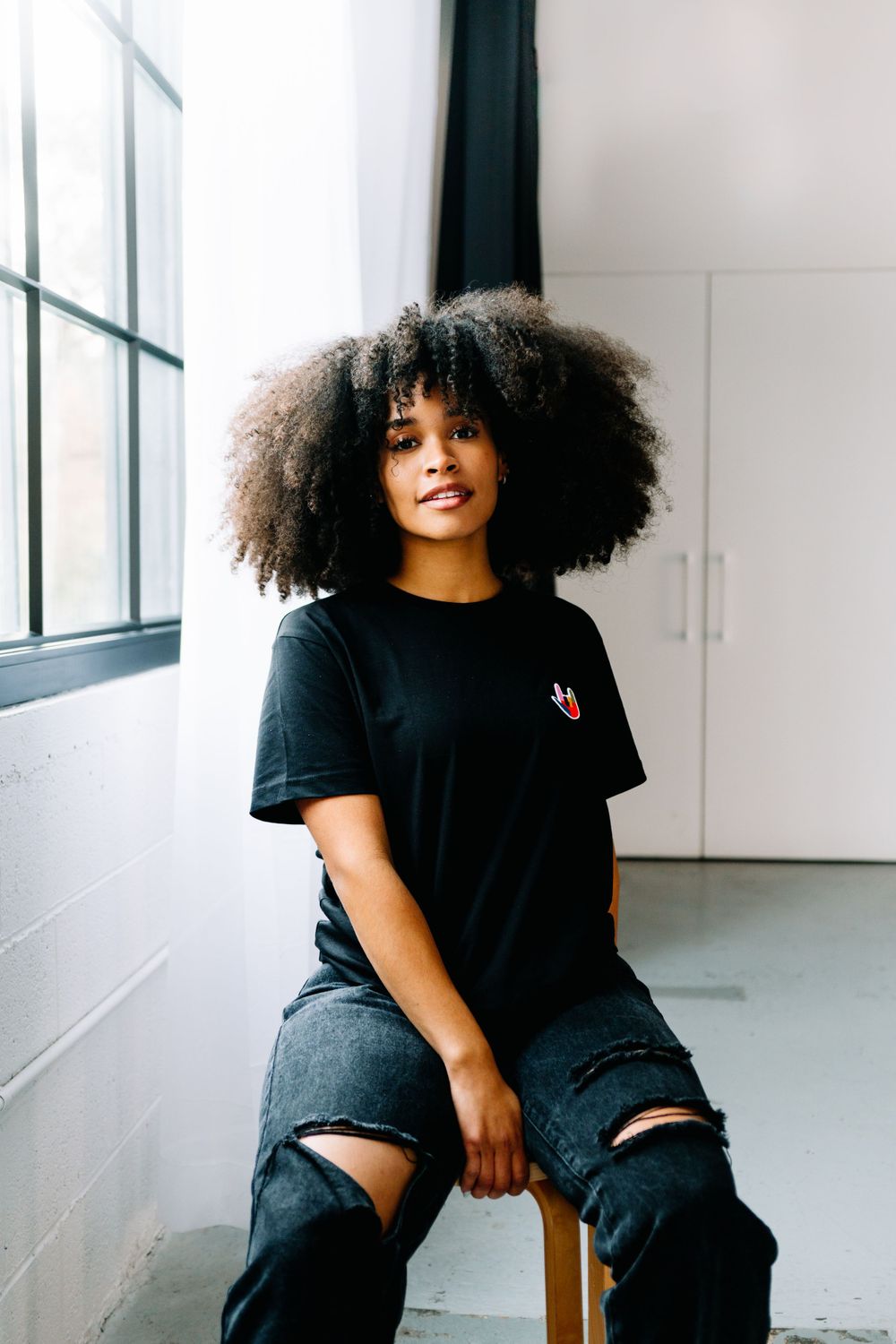 Model in black t-shirt sits casually on floor of minimalist white studio space.