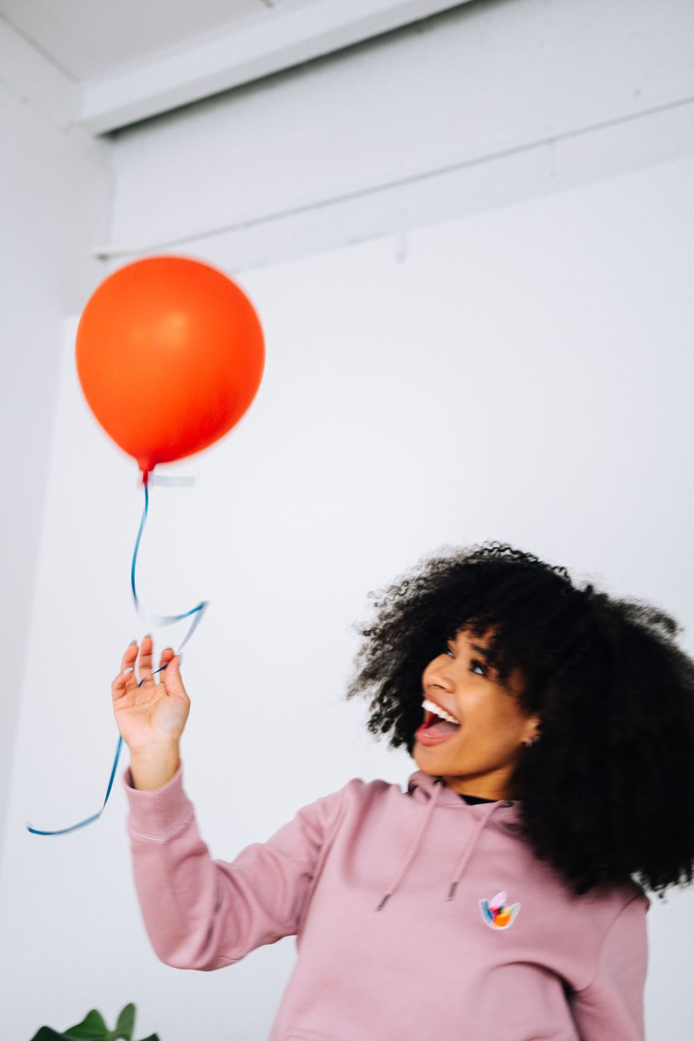 Model in pink hoodie playfully interacting with orange balloon in white studio setting.