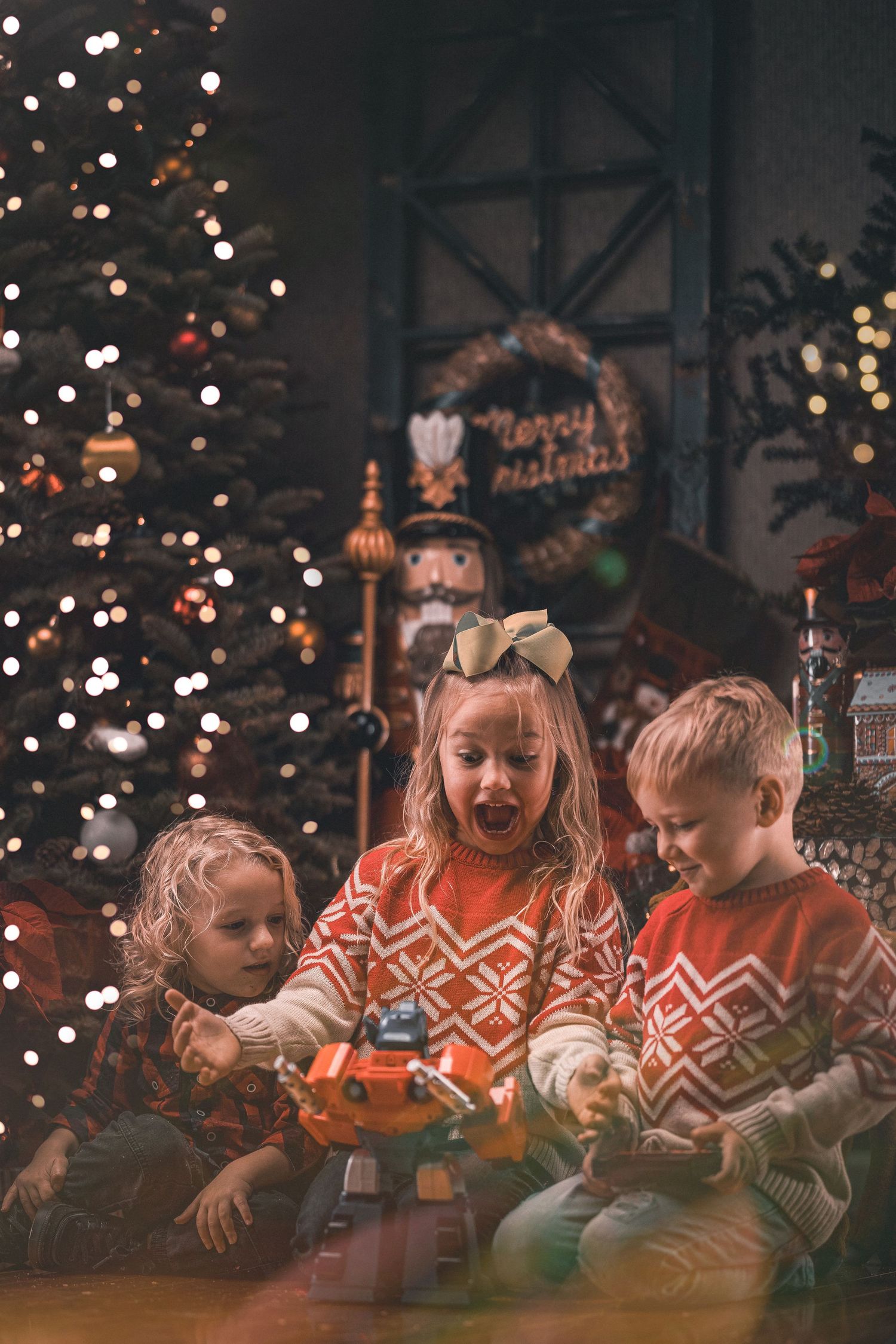 Three children in Christmas sweaters playing with toy train set near decorated holiday tree with lights.