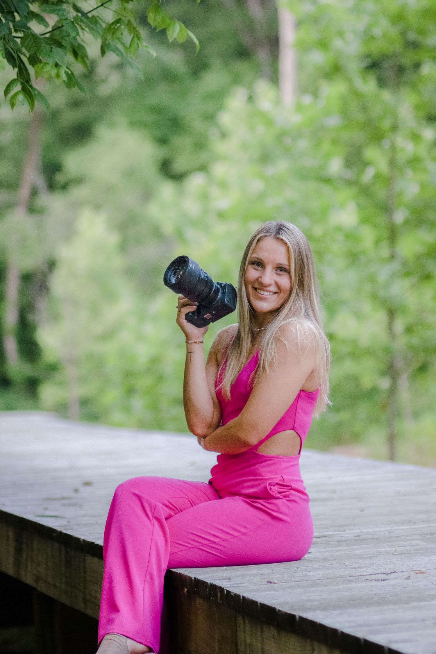Professional photographer wearing pink jumpsuit sits on wooden deck with camera in natural outdoor setting.