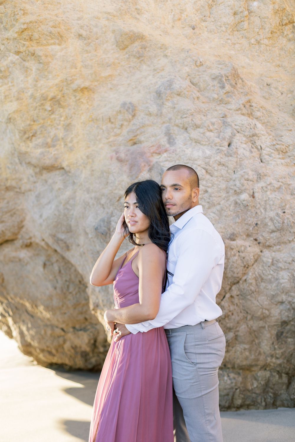 Elegant couple in formal attire poses against textured limestone cliff wall.