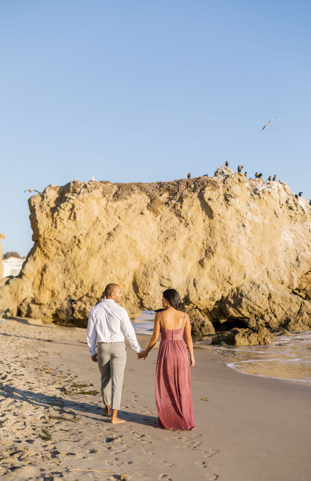 Couple walks hand in hand on beach with dramatic rock formation and seabirds in background.