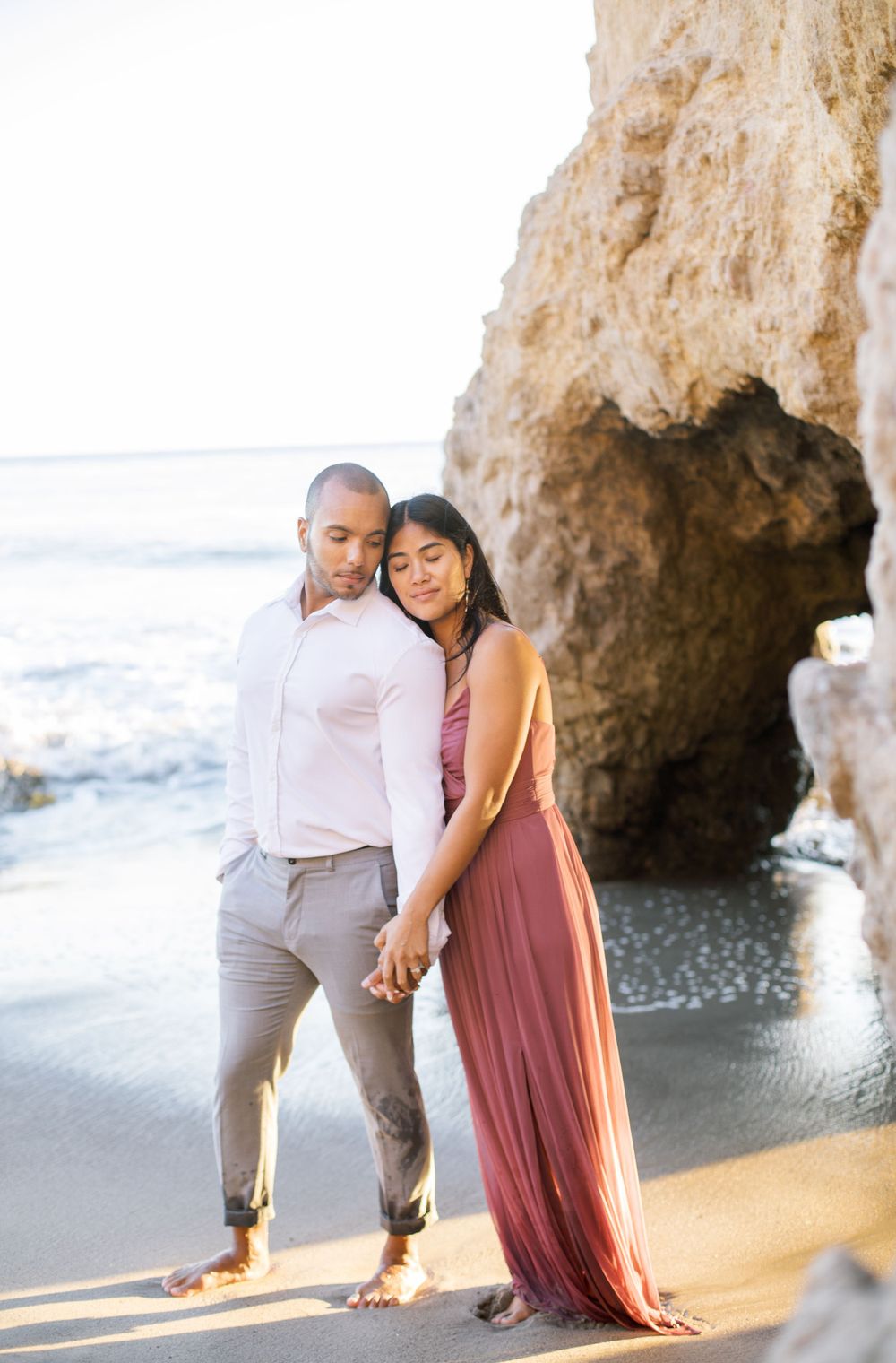 A couple in elegant attire poses together on a scenic beach with dramatic rock formations.