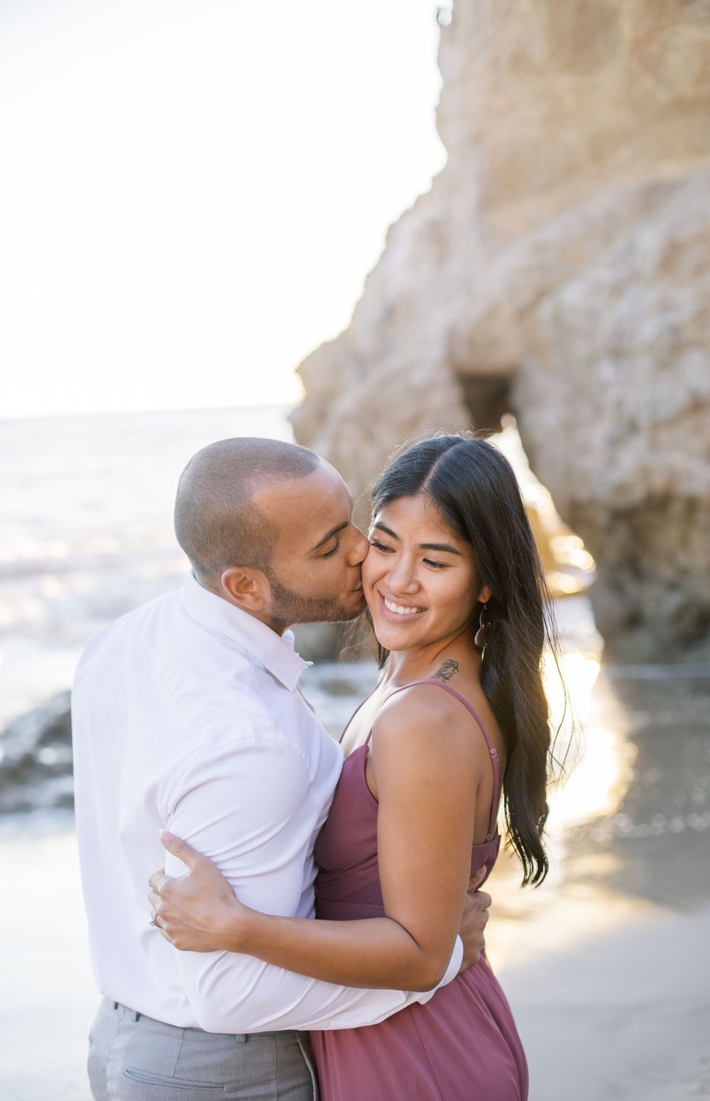 An intimate moment between a couple sharing a tender kiss on a beach with cliffs in the background.