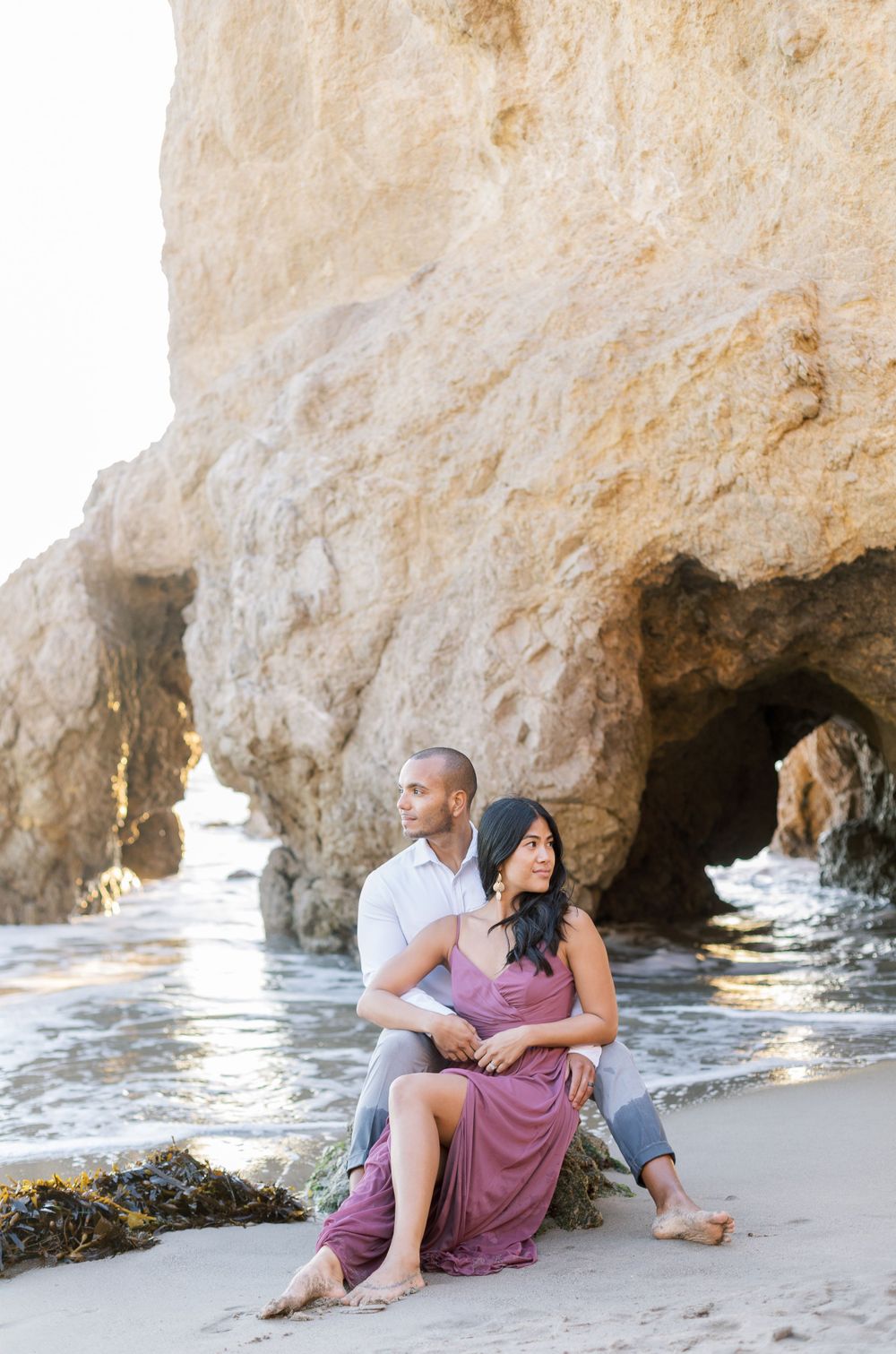 A couple poses together on a beach with dramatic rock formations and crashing waves.