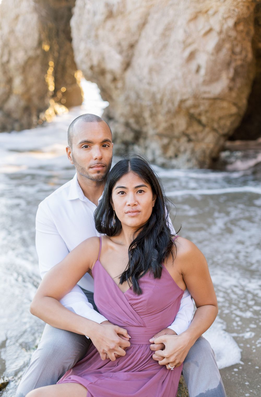 A romantic beach photoshoot of a couple embracing near rocky cliffs during golden hour.