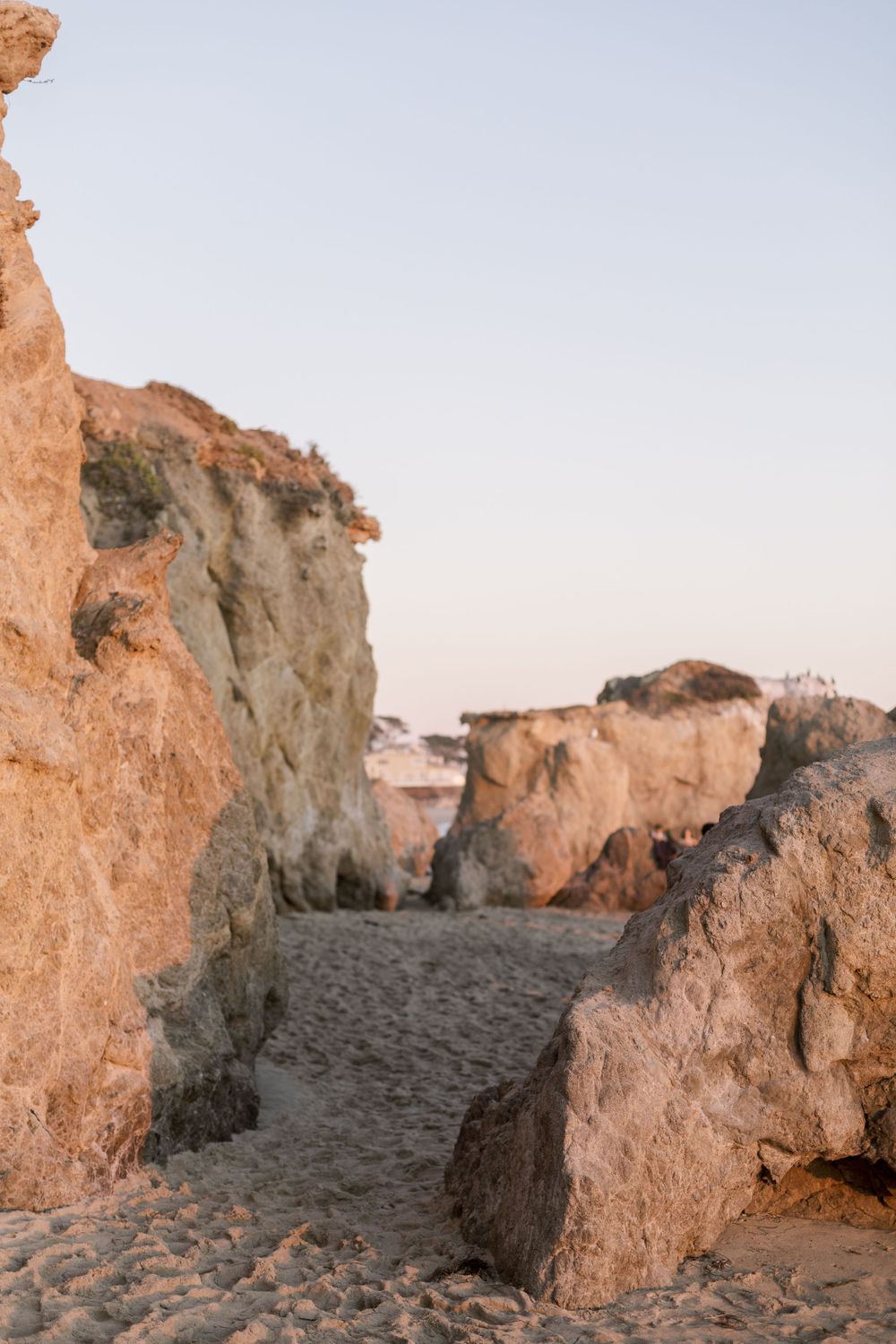 Rocky coastal cliffs and sandy pathway leading through natural stone formations at sunset.