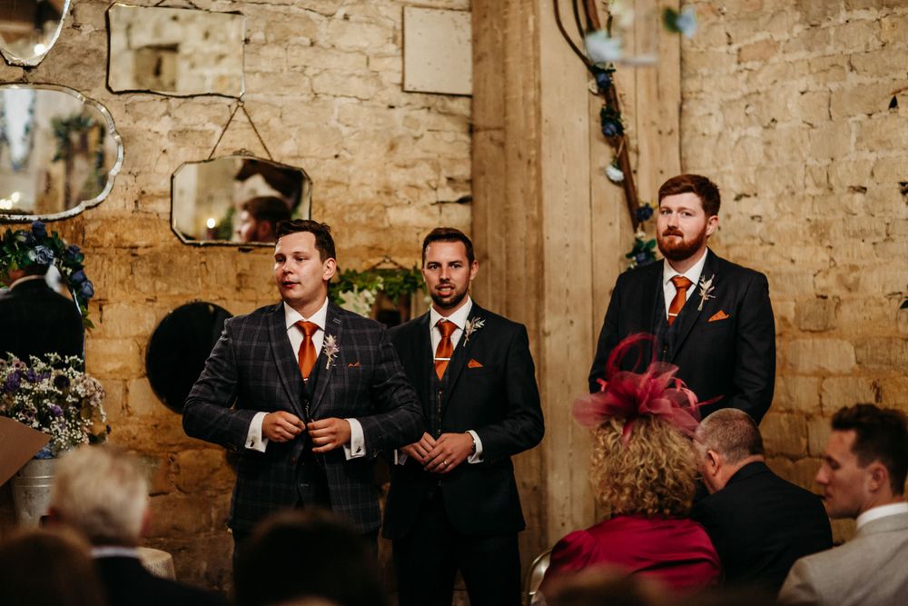 Formal wedding party members stand at attention during an indoor ceremony in a rustic stone venue.
