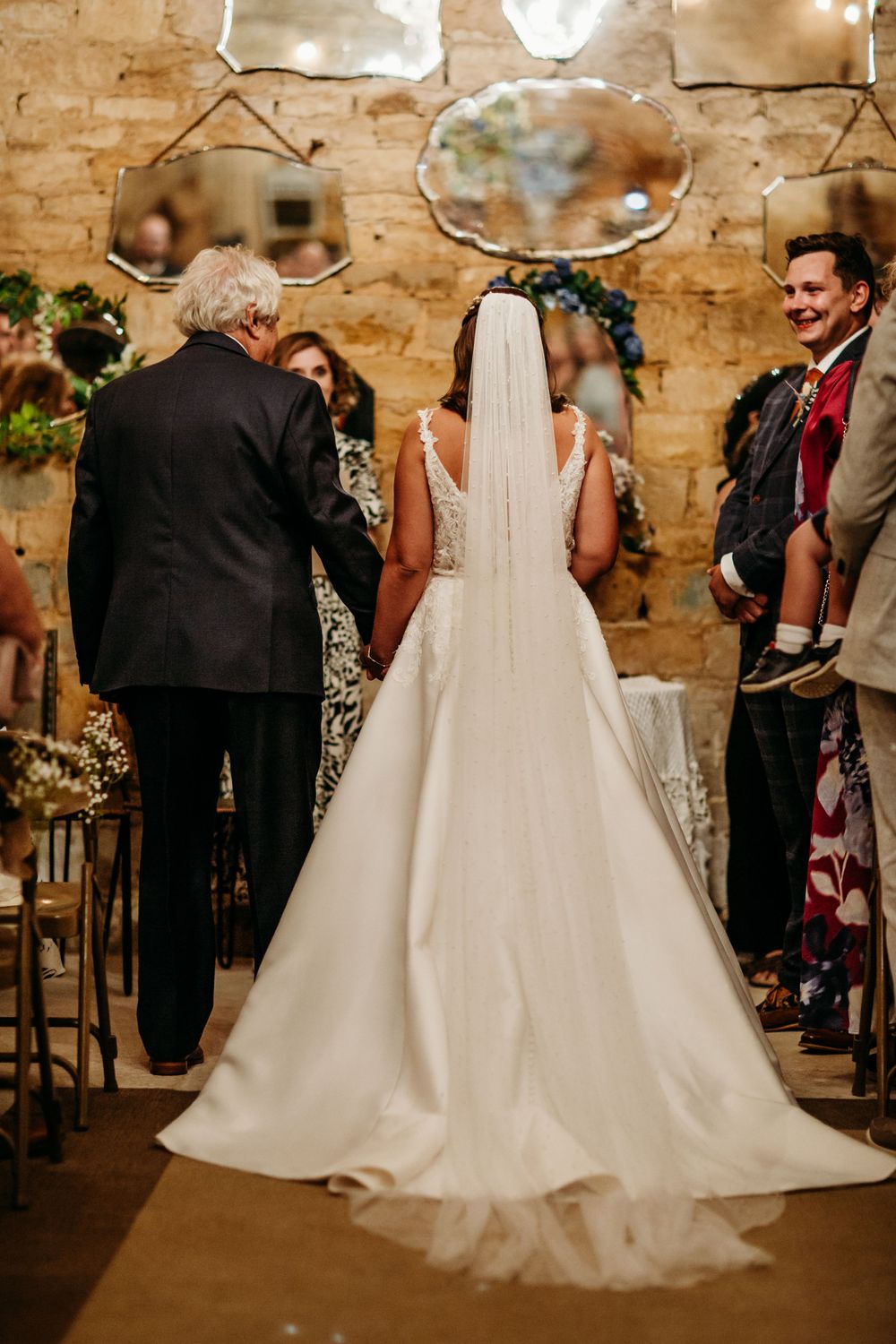Elegant bride in flowing white dress stands at rustic stone venue with vintage mirrors and chandeliers.