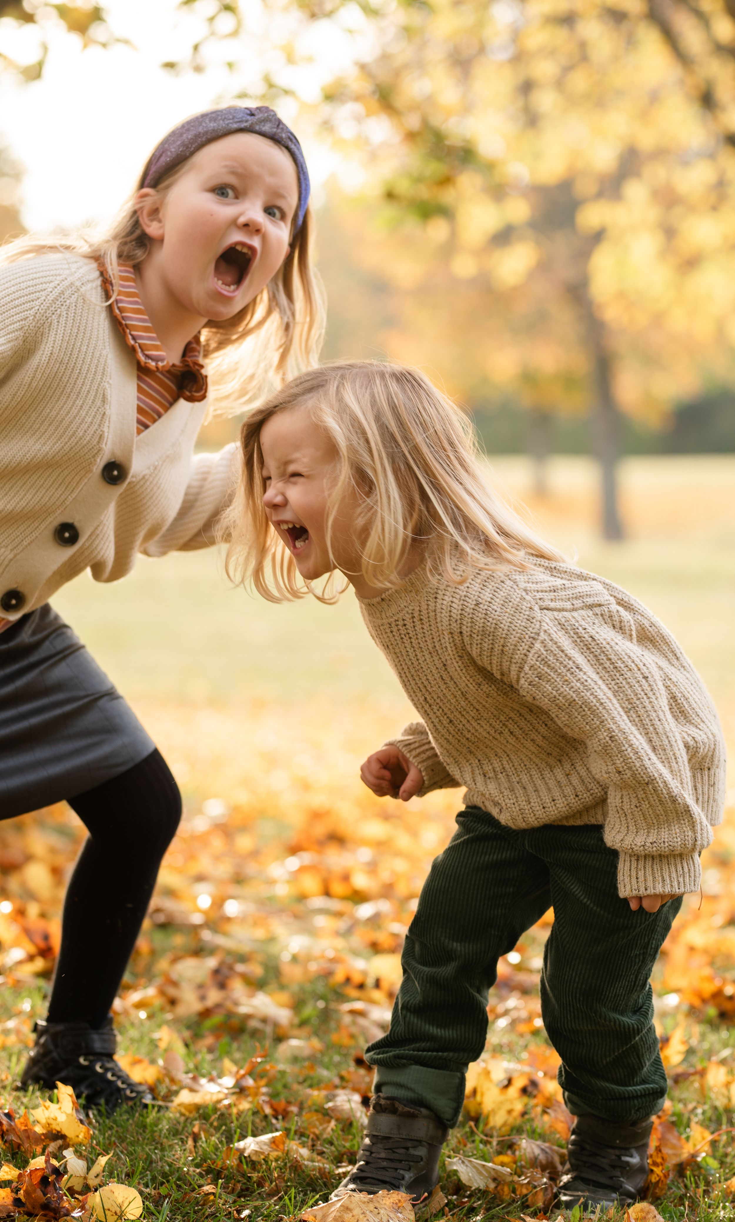 Two people in matching cream sweaters laughing and playing in fallen autumn leaves.