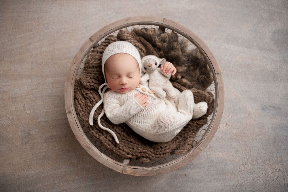 Newborn boy sleeping in a round basket, wearing a white knit sleeper and bonnet holding a tiny white lamb stuffed animal