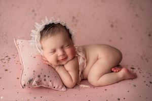 Newborn girl on her tummy, smiling in her sleep on a pink backdrop with gold stars and matching bonnet.