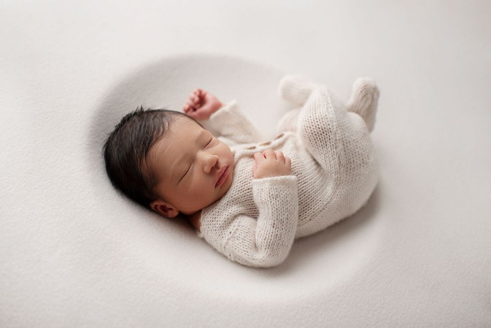 Sleeping newborn boy with lots of dark hair, curled up on his back, wearing a cream knit sleeper on a cream blanket.