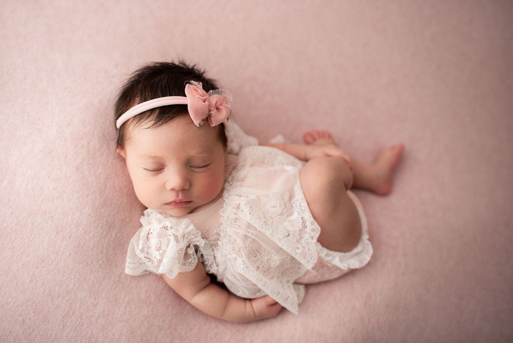 Sleeping newborn girl curled up on her back, wearing a white lace dress on a light pink fabric.