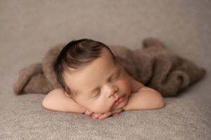 Sleeping newborn boy on his tummy with his hands under his chin on a light brown blanket.
