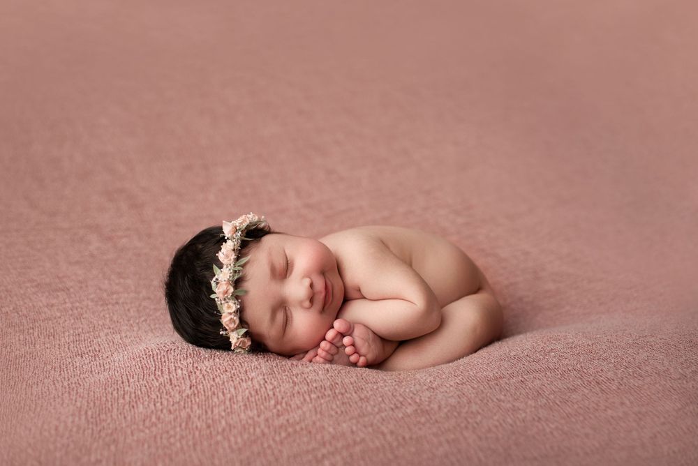 Sleeping newborn baby girl in a womb pose on dark rose pink fabric with a matching floral halo